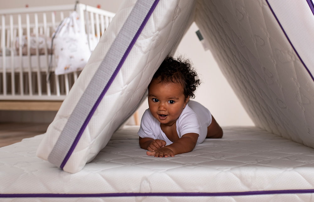 Baby unter aufgestellter Babymatratze mit Gitterbett im Hintergrund